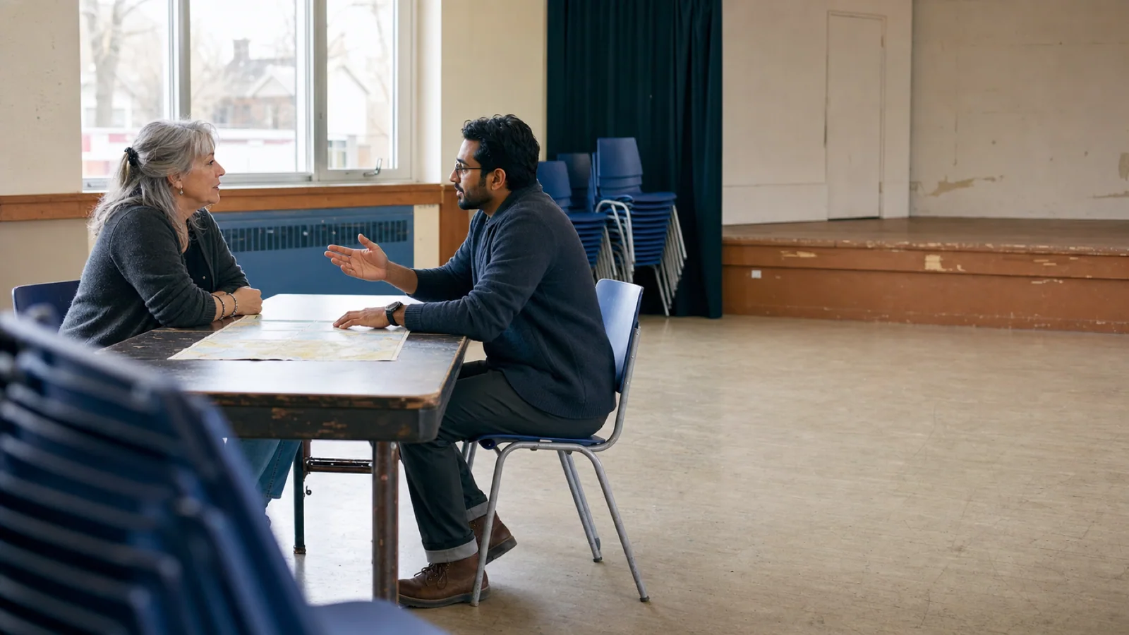 Deux adultes échangent autour d’un plan dans une salle communautaire.