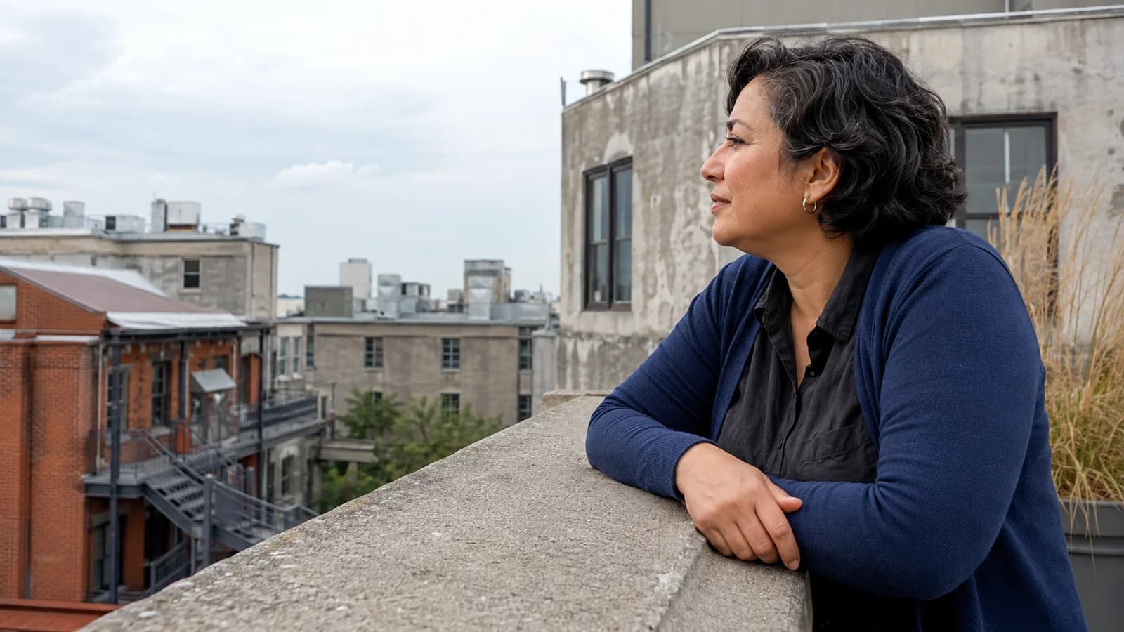 A woman in side profile wearing a blue cardigan pauses on a rooftop terrace overlooking brick buildings.
