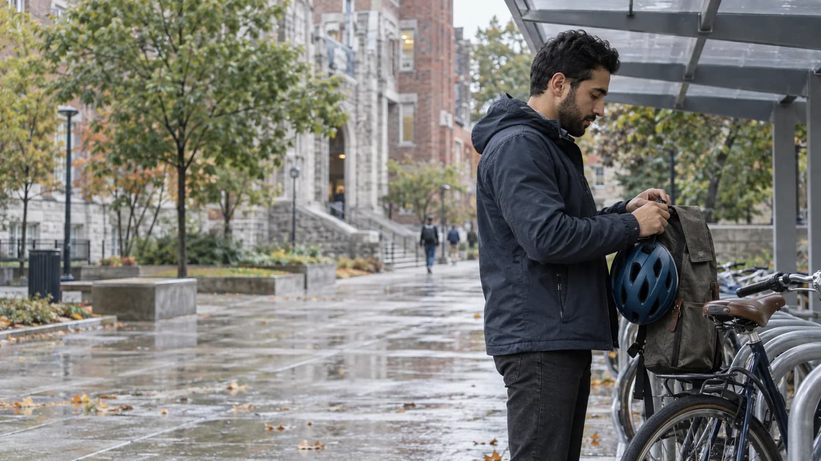 Outside a Montreal campus after rain, an adult student in side profile secures a blue bicycle helmet to a canvas bag under a covered bike rack.